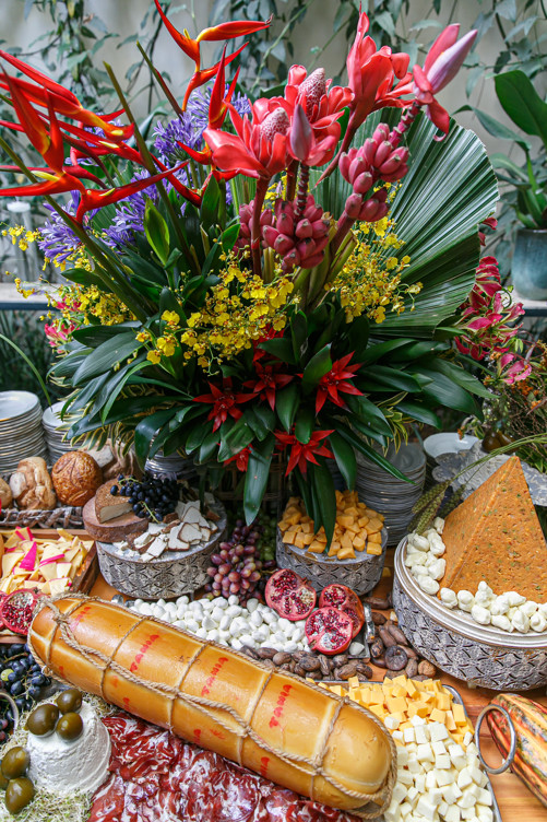 Arranjo tropical com helicônias, bastões-do-imperador e orquídeas sobre mesa de queijos e frios