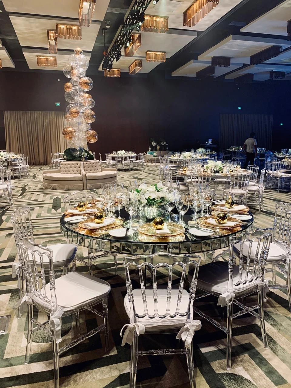 Jantar de Gala GS1 Brasil: mesa redonda espelhada com cadeiras transparentes, sousplats e esferas douradas, arranjo branco e torre de bolhas ao fundo